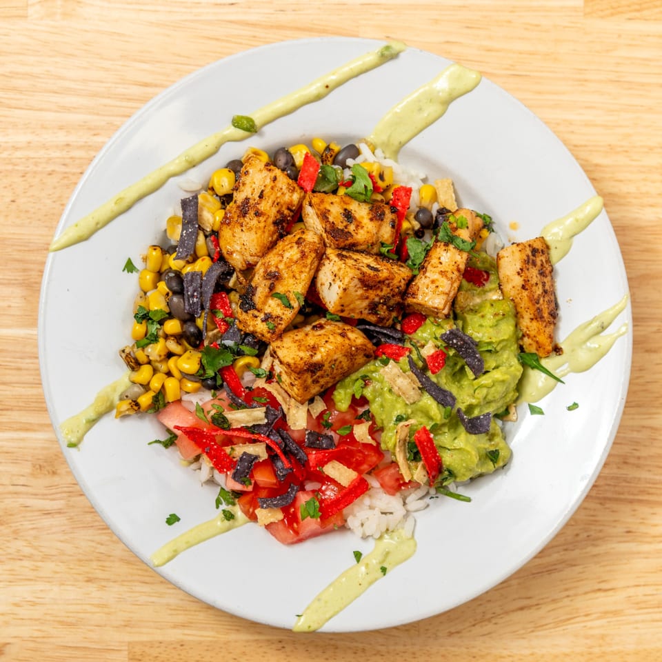 A colorful chicken bowl with rice, corn, black beans, guacamole, tomatoes, and tortilla strips.
