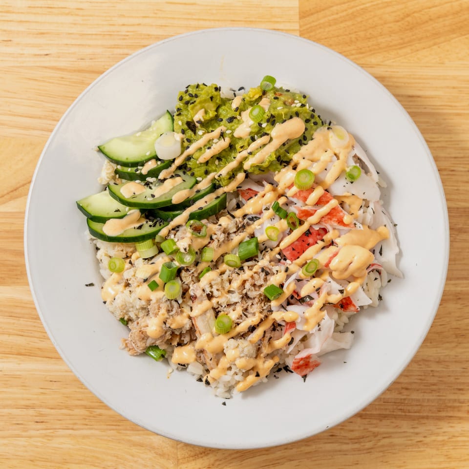 A poke bowl with rice, crab, avocado, cucumber, and a creamy sauce on a white plate.