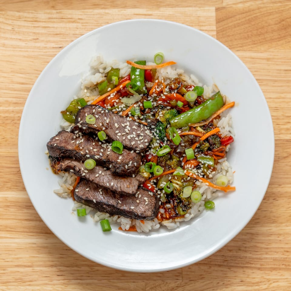 Steak and vegetable stir-fry with white rice, garnished with sesame seeds and green onions.